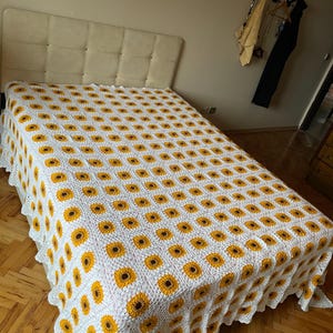 Puede incluir: Una manta de ganchillo blanca con un patr&oacute;n de girasol. La manta est&aacute; sobre una cama con una cabecera blanca.