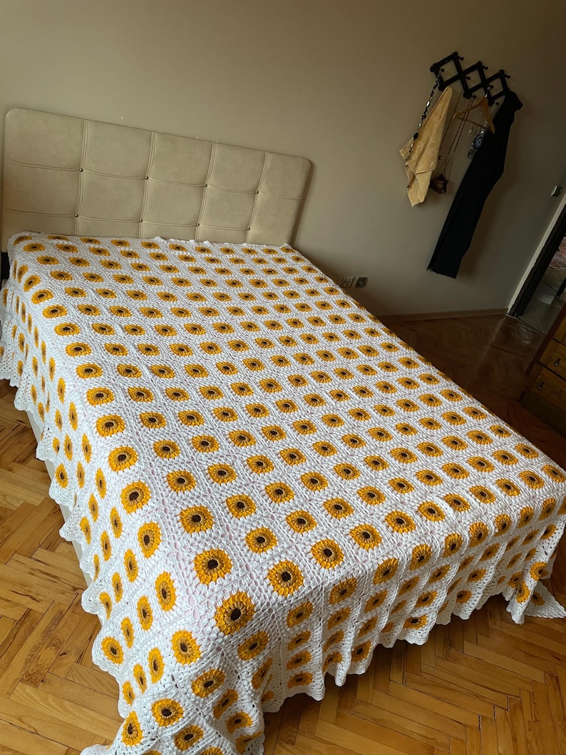 Puede incluir: Una manta de ganchillo blanca con un patr&oacute;n de girasol. La manta est&aacute; sobre una cama con una cabecera blanca.