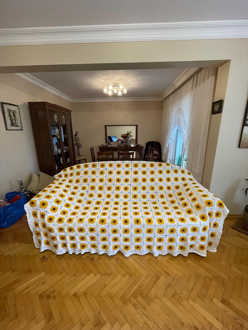Puede incluir: Una manta de crochet blanca con un patr&oacute;n de girasol. La manta est&aacute; colocada sobre un sof&aacute; en una sala de estar.