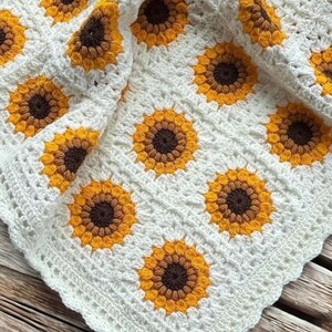 Puede incluir: Una manta de ganchillo blanca con un patr&oacute;n de girasol. La manta est&aacute; hecha con hilo amarillo, marr&oacute;n y blanco.