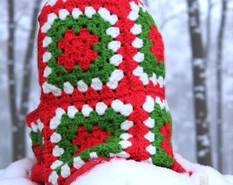 May include: A red, green, and white crocheted hood, likely a winter accessory. The hood features a granny square design with a repeating pattern of red, green, and white squares. The item is shown in a snowy outdoor setting.