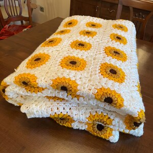 Puede incluir: Una manta blanca de ganchillo con un patr&oacute;n de girasol. La manta est&aacute; doblada y puesta sobre una mesa de madera.