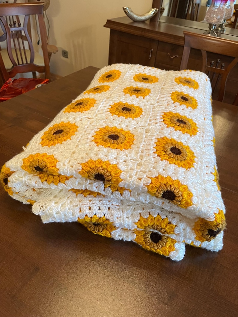 Puede incluir: Una manta blanca de ganchillo con un patr&oacute;n de girasol. La manta est&aacute; doblada y puesta sobre una mesa de madera.