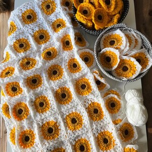Puede incluir: Una manta de ganchillo blanca con un patr&oacute;n de girasol. La manta est&aacute; hecha de cuadrados individuales con un centro amarillo y p&eacute;talos marrones. Hay dos cestas de cuadrados adicionales en la imagen.