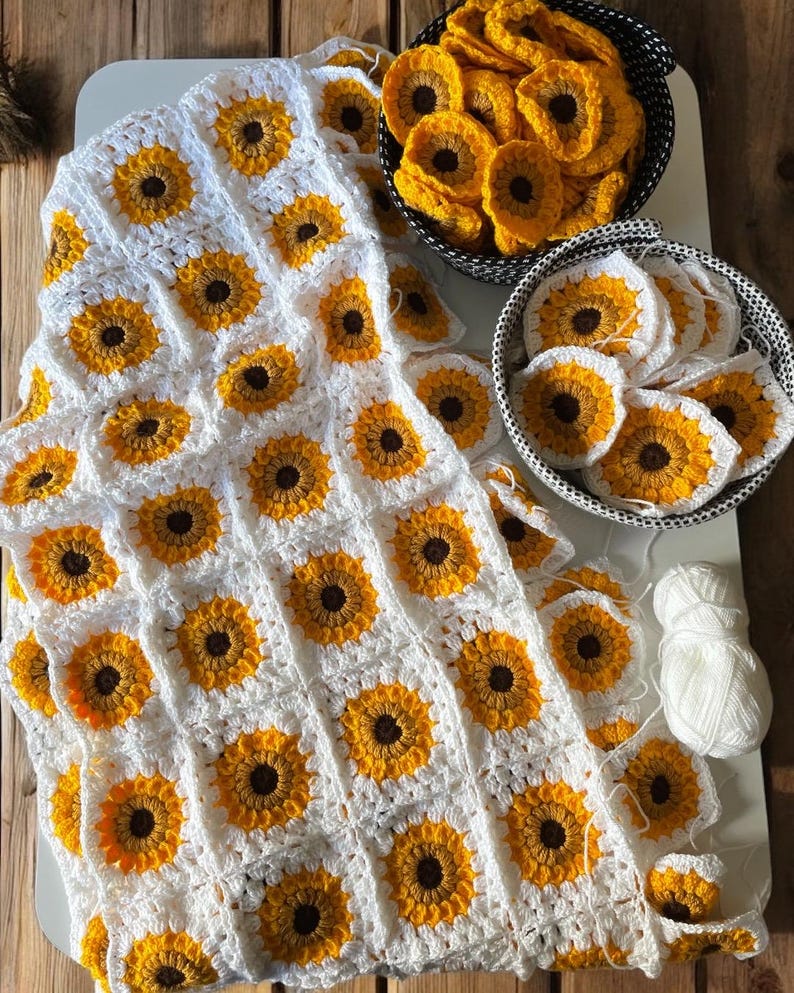 Puede incluir: Una manta de ganchillo blanca con un patr&oacute;n de girasol. La manta est&aacute; hecha de cuadrados individuales con un centro amarillo y p&eacute;talos marrones. Hay dos cestas de cuadrados adicionales en la imagen.