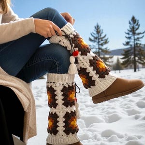 May include: Crocheted leg warmers in cream, brown, and yellow geometric patterns. The leg warmers have a tassel and pom-pom detail. The person is wearing blue jeans and brown boots. The background is a snowy landscape.