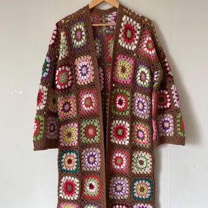 Könnte beinhalten: Ein langer brauner Häkelcardigan mit einem bunten Granny-Square-Muster. Der Cardigan hat einen V-Ausschnitt und lange Ärmel.