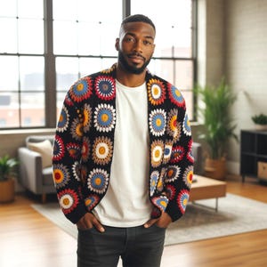 May include: A black crochet cardigan with colorful floral patterns in red, blue, yellow, and white. The open-front sweater is worn over a white t-shirt and dark pants. The cardigan's design features a unique, handmade aesthetic.
