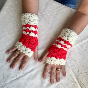 Könnte beinhalten: Ein Paar handgefertigte Fingerhandschuhe aus Häkelarbeit. Die Handschuhe haben eine cremefarbene Manschette und ein rotes und cremefarbenes Muschelmuster. Die Handschuhe sind so konzipiert, dass sie das Handgelenk und einen Teil der Hand bedecken.