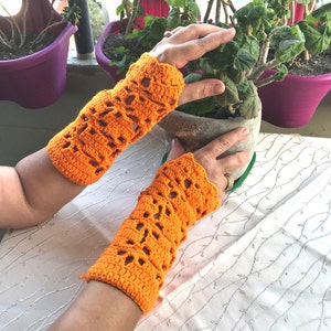 Könnte beinhalten: Ein Paar leuchtend orangefarbene Armstulpen aus Häkelarbeit mit einem dekorativen Lochmuster. Die handgefertigten Fingerhandschuhe werden auf einer weißen Oberfläche gezeigt, mit einer Pflanze und lila Blumentöpfen im Hintergrund.