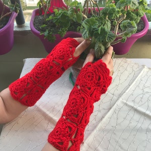 Könnte beinhalten: Ein Paar leuchtend rote, offene Finger-Häkelhandschuhe. Die Handschuhe weisen ein detailliertes Blumenmuster auf und sind so konzipiert, dass sie die Hand und den Unterarm bedecken. Die Häkelarbeit ist aufwendig, mit offenen Stellen, die ein zartes Aussehen erzeugen. Die Handschuhe sind wahrscheinlich handgefertigt.
