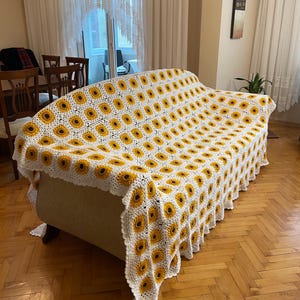 Puede incluir: Una manta de crochet blanca con un patr&oacute;n de girasol en amarillo y marr&oacute;n. La manta est&aacute; drapeada sobre un sof&aacute; blanco.