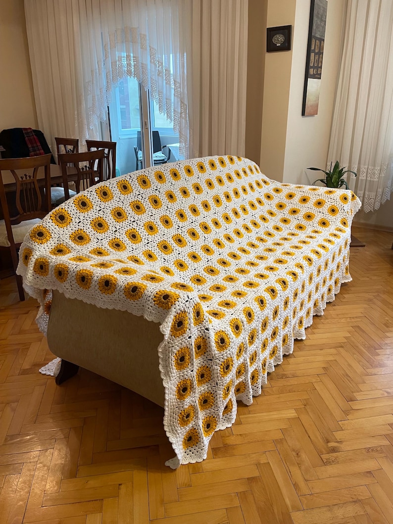Puede incluir: Una manta de crochet blanca con un patr&oacute;n de girasol en amarillo y marr&oacute;n. La manta est&aacute; drapeada sobre un sof&aacute; blanco.