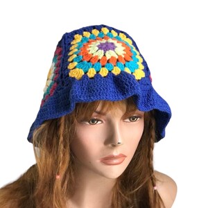 May include: A blue crochet bucket hat with a colorful square pattern on the crown. The square pattern features orange, yellow, teal, and purple. The hat has a ruffled brim.