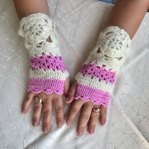 Könnte beinhalten: Ein Paar handgefertigte Fingerhandschuhe aus Häkelarbeit. Die Handschuhe haben ein cremefarbenes Blumendesign am Handgelenk und ein rosa-cremefarbenes Streifenmuster. Die Handschuhe bedecken Hand und Handgelenk, wobei die Finger frei bleiben.