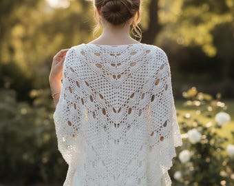 May include: A white, crocheted shawl with a detailed openwork design. The shawl is draped over the shoulders, with a focus on the intricate patterns and textures of the handmade garment. The background is a soft, blurred outdoor scene.