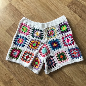 Könnte beinhalten: Weiße Häkelshorts mit einem bunten Granny-Square-Muster. Die Shorts haben einen Kordelzugbund und sind aus verschiedenen Farben gefertigt, darunter Pink, Blau, Grün, Gelb und Lila.