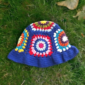 May include: A blue crochet bucket hat with a colorful granny square design. The hat features a wide brim and is made with various colors including red, yellow, orange, and white. The hat is sitting on a bed of green grass.