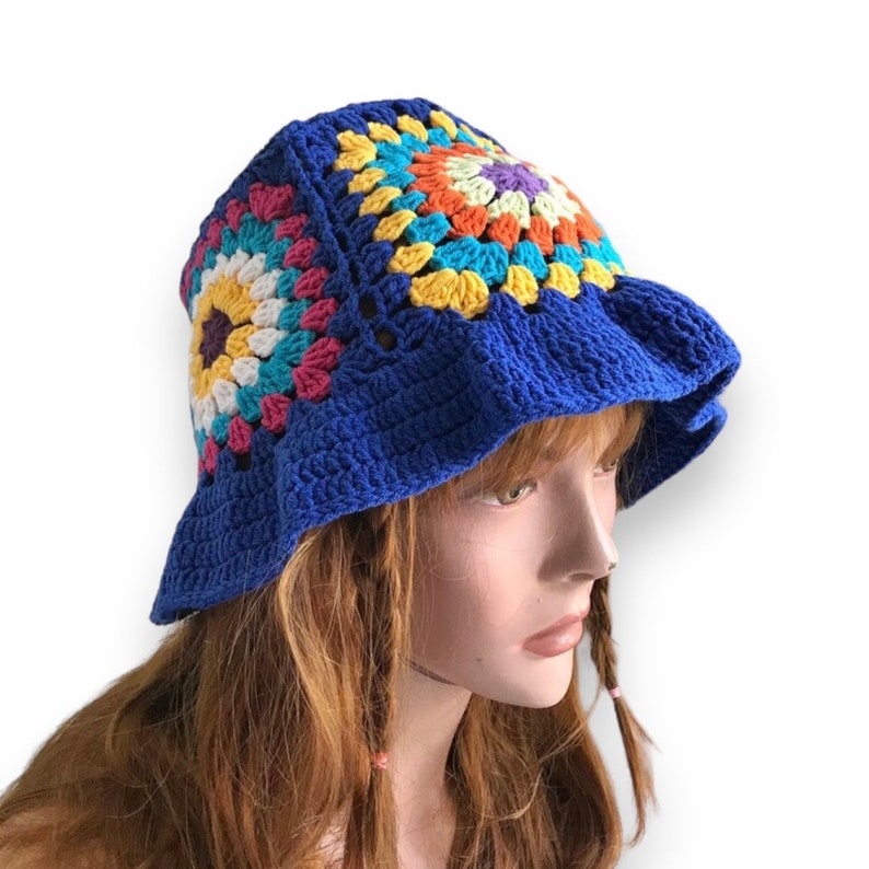 May include: A blue crochet bucket hat with a colorful floral design. The hat features a wide brim and is made with a granny square pattern in shades of yellow, pink, turquoise, and orange. The hat is displayed on a mannequin head.