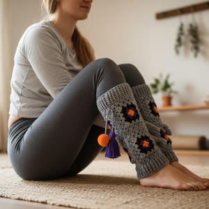 May include: Gray crochet leg warmers with orange and black floral designs and purple and orange tassels. The leg warmers are worn with gray leggings. The image is taken indoors on a woven mat.