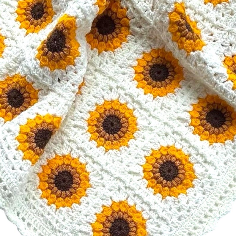 Puede incluir: Manta de beb&eacute; de crochet blanco con un patr&oacute;n de girasol. La manta est&aacute; hecha con hilo amarillo, marr&oacute;n y blanco.