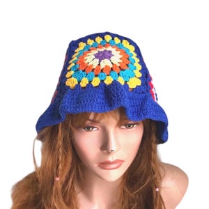 May include: A blue crochet bucket hat with a colorful square design on top. The square features orange, yellow, blue, and purple accents. The hat has a ruffled brim and is made of yarn.
