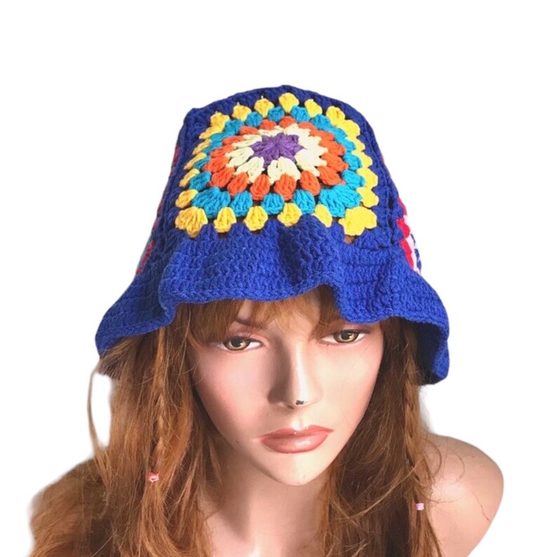 May include: A blue crochet bucket hat with a colorful square design on top. The square features orange, yellow, blue, and purple accents. The hat has a ruffled brim and is made of yarn.