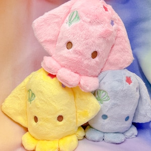 Op de afbeelding: Drie pluche octopus speelgoed in roze, geel en blauw. Elke octopus heeft geborduurde details, waaronder schelpen en sterren.