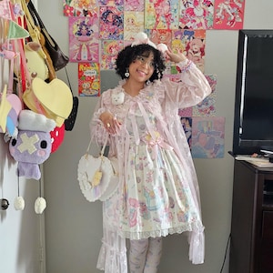 May include: A light pink Lolita dress with a whimsical print, paired with matching leggings and shoes. The outfit includes a heart-shaped purse and a sheer, ruffled cape. The background features colorful anime art and various hanging bags.