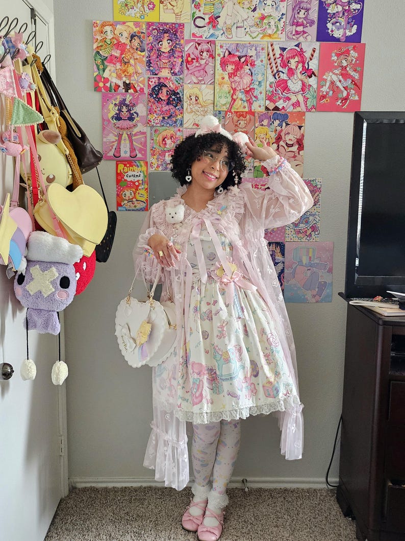 May include: A light pink Lolita dress with a whimsical print, paired with matching leggings and shoes. The outfit includes a heart-shaped purse and a sheer, ruffled cape. The background features colorful anime art and various hanging bags.