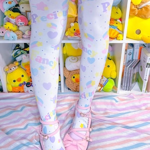 May include: White leggings with a pastel pattern of hearts, rainbows, and the word "Fancy". The leggings are paired with pink Mary Jane shoes with bows. The outfit is set against a backdrop of stuffed animals and a striped bedspread.