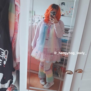 May include: A person wearing a light pink outfit with a rainbow-colored tulle overlay and matching leg warmers. The outfit includes a dress and a long-sleeved top. The person is reflected in a mirror. The text "slim" is visible on the outfit.