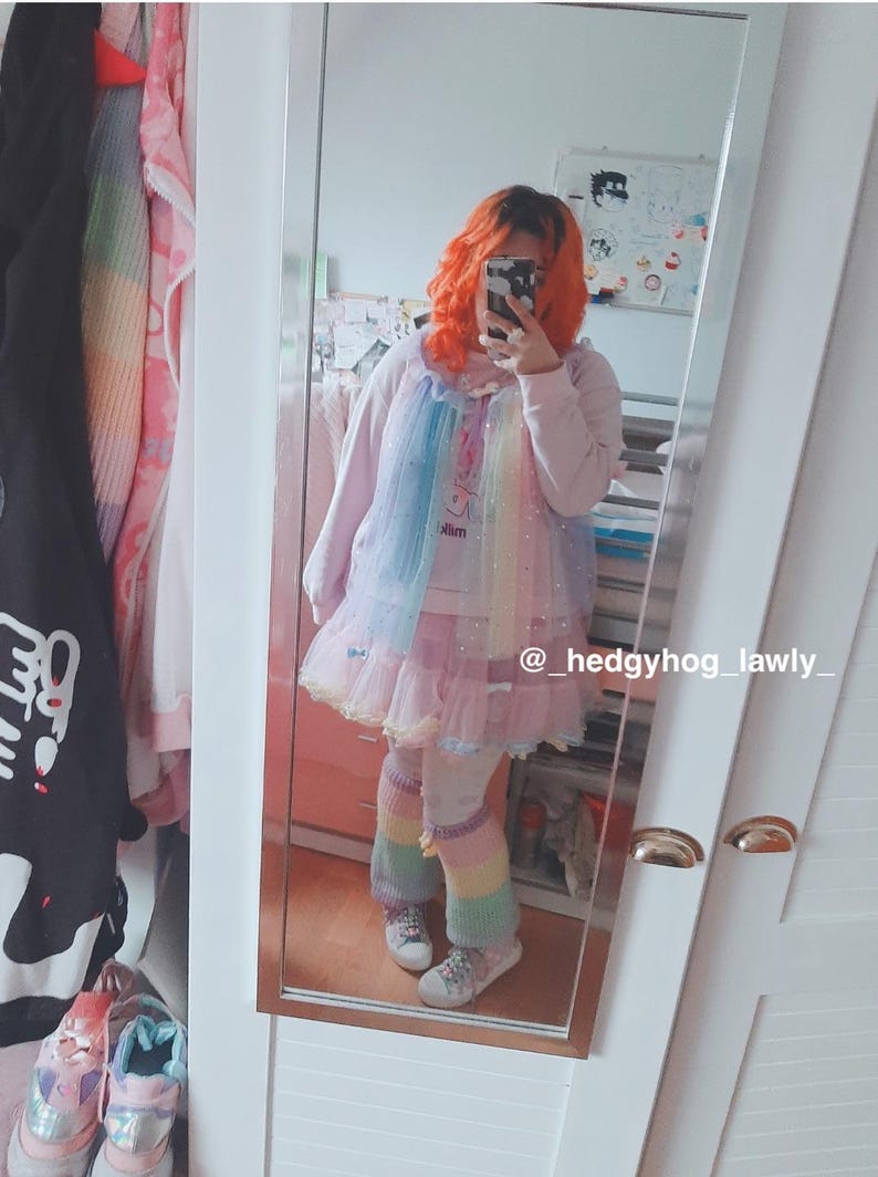 May include: A person wearing a light pink outfit with a rainbow-colored tulle overlay and matching leg warmers. The outfit includes a dress and a long-sleeved top. The person is reflected in a mirror. The text "slim" is visible on the outfit.