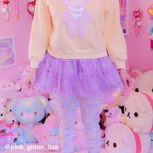 May include: A pastel yellow sweatshirt with a lavender teddy bear applique, paired with a lavender tulle skirt and patterned leggings. The outfit is completed with yellow sneakers. The background is a pink room with plush toys.