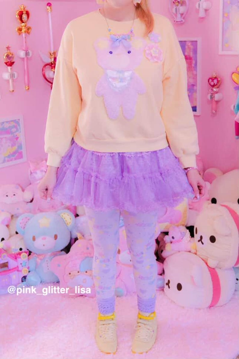 May include: A pastel yellow sweatshirt with a lavender teddy bear applique, paired with a lavender tulle skirt and patterned leggings. The outfit is completed with yellow sneakers. The background is a pink room with plush toys.