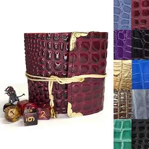 May include: A burgundy faux leather dice rolling tower with a crocodile pattern and gold accents. The tower is tied with a gold rope and has a skull charm. There are five dice on the table in front of the tower.