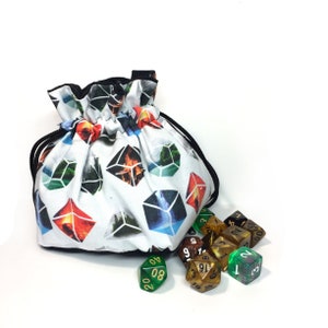 Può includere: Una borsa a cordoncino bianca con un cordoncino nero e un motivo geometrico colorato. La borsa è piena di dadi, alcuni sono verdi, alcuni sono dorati e alcuni sono marroni.