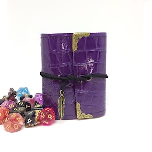 May include: Purple leather dice bag with a crocodile pattern, a black cord closure, and a gold feather charm. The bag is closed and sitting on a white surface with a pile of colourful dice in front of it.