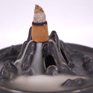Op de afbeelding: Zwarte keramische wierookbrander in de vorm van een berg met een enkele wierookkegel die erboven brandt. De rook van de kegel stroomt de berg af.