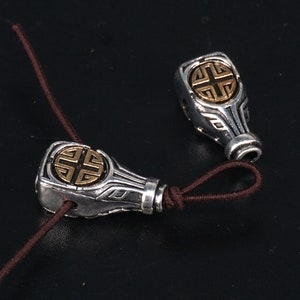 May include: Two silver and gold charm beads with a knot design and a square symbol in the centre. The beads are strung on a brown cord.