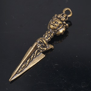 May include: A gold-colored metal pendant in the shape of a dagger with a detailed design. The dagger has a face on the top and a loop for hanging.
