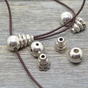 Könnte beinhalten: Ein Set aus silbernen Metallperlen und Endkappen für die Schmuckherstellung. Die Perlen sind rund und haben eine glatte Oberfläche. Die Endkappen sind kegelförmig und haben ein dekoratives Design.