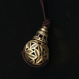 Op de afbeelding: Een gouden metalen hanger met een decoratief, ingewikkeld ontwerp. De hanger is bevestigd aan een bruin koord.