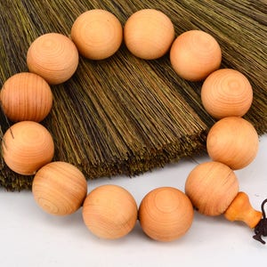 May include: A wooden beaded necklace with a brown tassel. The beads are round and have a natural wood color. The necklace is lying on a brown, woven surface.
