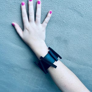 May include: A black geometric bracelet with a wide band and multiple angular shapes. The bracelet is worn on a person's wrist.