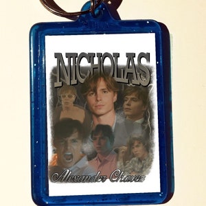 May include: Blue plastic keychain with a white background featuring a collage of images of a young man with the text "NICHOLAS Alexander Chavez" printed in black.