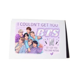 May include: A white greeting card featuring a colorful illustration of the seven members of the music group BTS. The text reads I COULDN'T GET YOU BTS SO I GOT YOU THIS CARD INSTEAD. The design includes two light sticks, a logo, and a purple heart.