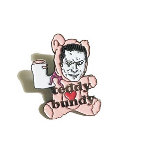 May include: A pink teddy bear pin with a cartoon face of a man wearing glasses. The bear is holding a white object in its left hand and the text "teddy ♥ bundy" is printed on the bear's chest.