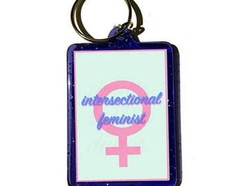 May include: A blue keychain with a pink female symbol and the text "intersectional feminist" on a light blue background.