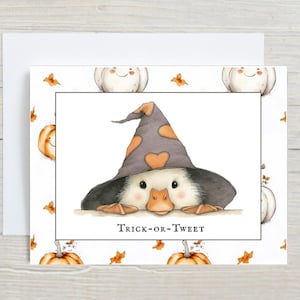 Pu&ograve; includere: Biglietto di auguri di Halloween con un'anatra dei cartoni animati con un cappello da strega con forme di cuore arancioni. Il biglietto ha uno sfondo bianco con zucche e foglie. Il testo "Trick-or-Tweet" &egrave; sotto l'anatra.